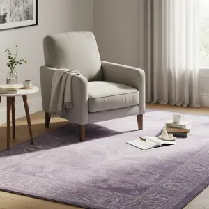 Cozy reading nook with a comfortable armchair, a soft lavender patterned rug, a side table with books, and natural light from a window, creating a tranquil and inviting atmosphere.